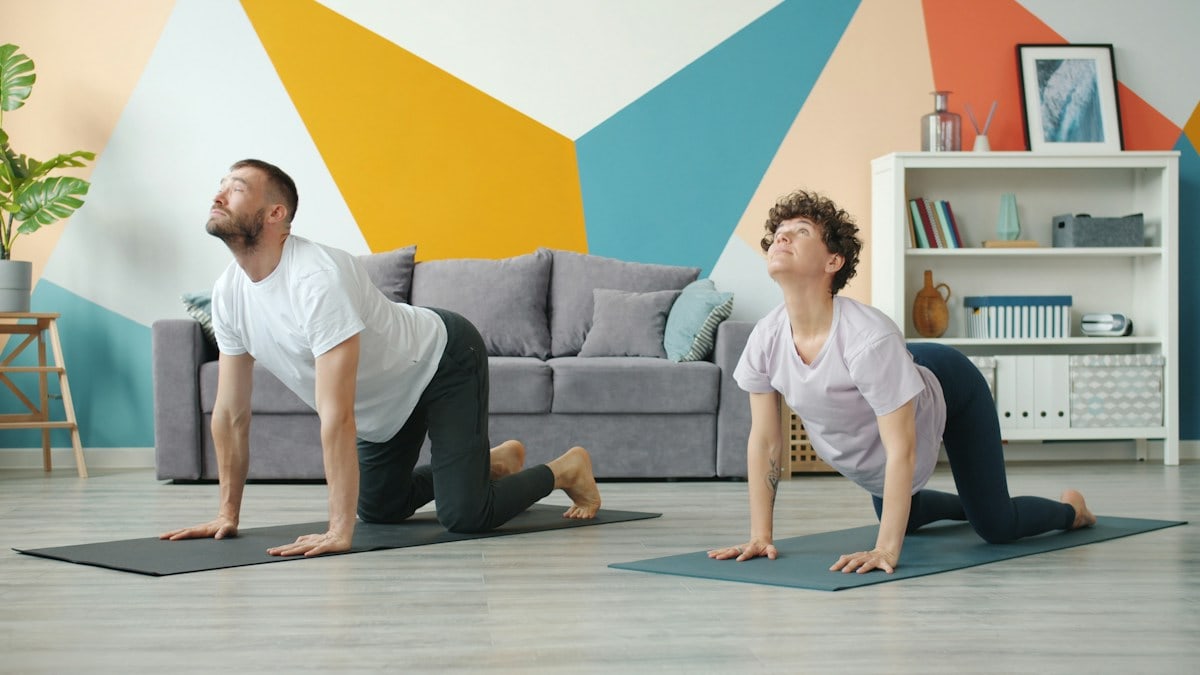 Slow motion of young attractive husband and wife doing yoga in apartment together caring for health training body. Cozy room is visible in background.